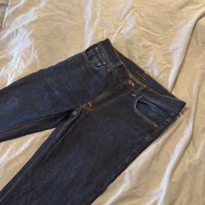 Nudie lean dean 30/32  - Snygga mörkblå jeans från Nudie Jeans med klassiska orange sömmar och femficksdesign. Jeansen är slimfit i modellen och har en stilren look med Nudie-logga på bakfickan. Perfekta för dig som gillar en tidlös och clean stil.