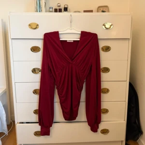 Vinröd omlottopp med v-ringning - Säljer en vinröd omlottopp med djup v-ringning och långa ärmar. Toppen har snygga rynkade detaljer framtill och är tillverkad i ett mjukt, stretchigt material. Perfekt för dig som vill ha en trendig och bekväm look.