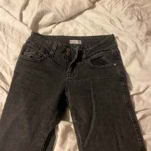 Svarta jeansbyxor från Perfect Jeans - Svarta jeansbyxor från Perfect Jeans med hög midja och raka ben. Byxorna har dubbla knappar i midjan, klassiska femficksdetaljer och en bakficka med lock och knapp. Materialet är jeans och färgen är mörksvart.