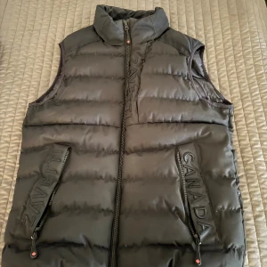 Svart dunväst från Zavetti Canada - Svart, quiltad dunväst från Zavetti Canada med hög krage och dragkedja framtill. Västen har broderad logga på ryggen, diskreta logotyper på fickorna och insidan, samt två praktiska fickor med dragkedja. Perfekt för lager-på-lager under kyliga dagar.