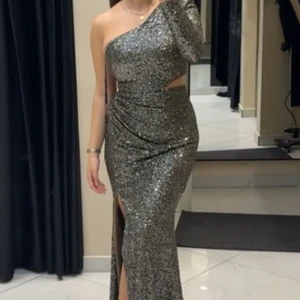 Glittrig silver långklänning oneshoulder - Säljer en maxad långklänning i glittrigt silver med en ärm och snygg oneshoulder-design. Klänningen har cut-out vid midjan, slits framtill och sitter tight för en riktigt elegant look. Perfekt för dig som vill sticka ut på bal eller fest! Använd enbart vid 1 tillfälle.