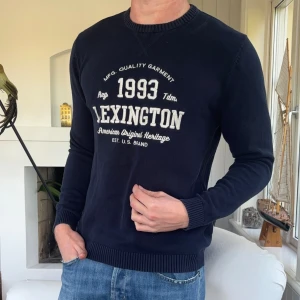 Mörkblå Lexington tröja med tryck - Snygg mörkblå tröja från Lexington med vitt tryck framtill och klassisk rund hals. Tröjan är i bomull och har ribbade muddar vid ärmslut och nederkant. Perfekt för dig som gillar stilren och sportig look.