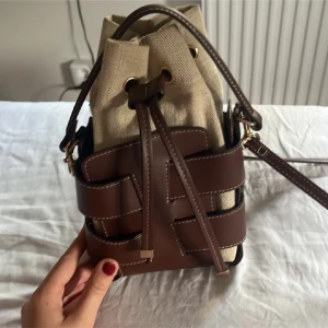 Brun och beige bucket bag med snörning - Snygg bucket bag i beige canvas och brunt skinn med coola detaljer och snörning upptill. Väskan har axelrem och guldiga metalldetaljer. Perfekt storlek för det viktigaste och ger en trendig touch till din outfit.