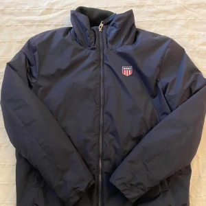 Marinblå vindjacka från GANT - Snygg marinblå vindjacka från GANT med dragkedja framtill, hög krage och broderad logga på bröstet. Jackan har elastisk nederkant med GANT-tryck och är tillverkad i polyester. Perfekt för dig som gillar stilren och sportig look. Perfekt nu till hösten och väldigt stilren om du frågar mig och den kan göra dina outfits felfria😉 Tveka inte att kontakta vid minsta fundering eller fråga!👊 