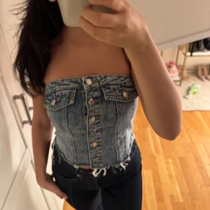 Jeans topp - Säljer en snygg jeans topp  med silverfärgade knappar. Toppen är axelbandslös och har en cool denim-look med fickdetaljer. Perfekt för dig som vill sticka ut med en unik och trendig stil.
