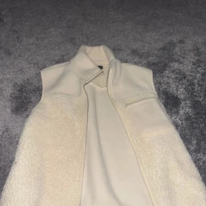 Beige fleeceväst med hög krage - Mysig beige fleeceväst med hög krage och en bröstficka. Västen har en enkel och stilren design med mjukt teddyfoder på framsidan. Perfekt att slänga över en hoodie eller tröja för extra värme.