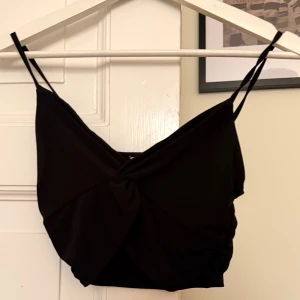 Svart vriden topp från Pull&Bear - Svart croppad vriden topp från Pull&Bear med tunna axelband och snygg omlottdesign framtill. Toppen har en tight passform och är tillverkad i mjuk polyester, perfekt för att lyfta vilken outfit som helst.