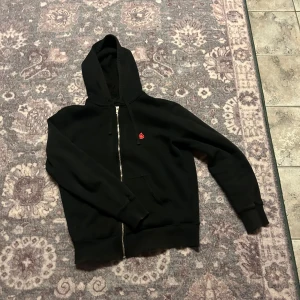 Svart hoodie från Bloodline med dragkedja - Svart hoodie från Bloodline med hel dragkedja framtill och huva med snörning. Tröjan har en liten röd broderad detalj på bröstet och praktiska fickor. Perfekt för lager-på-lager och chill dagar.