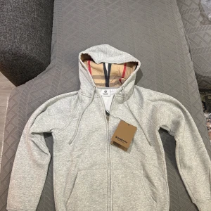 Grå hoodie från Burberry med dragkedja - Snygg grå hoodie från Burberry med klassiskt rutigt foder i huvan. Tröjan har dragkedja framtill, snörning i huvan och två fickor. Perfekt för dig som vill ha en stilren och exklusiv look med en touch av lyx. Desutom helt ny och aldrig använd, köpte fel storlek