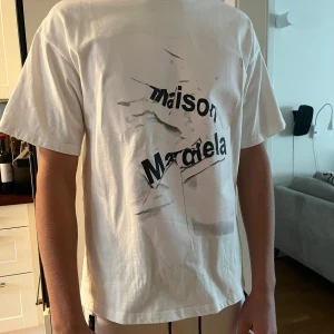 Maison Margiela vit t-shirt med tryck - Vit t-shirt från Maison Margiela med svart grafiskt tryck på bröstet. Oversized fit med korta ärmar och rund halsringning. Tillverkad i mjuk bomull för en skön känsla. Perfekt för dig som gillar statement-plagg och streetstyle. Tröjan har änglars fåtal gånger men har en liten grå fläck samt en sömm som har lossnat men är lätt att sy på.