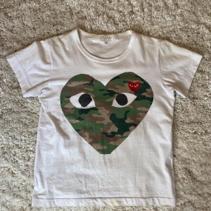 Vit Comme des Garçons Play t-shirt - Säljer en vit t-shirt från Comme des Garçons Play med stort hjärta i camouflage på bröstet och broderat rött hjärta med ögon. Klassisk crewneck och korta ärmar. Hör av för frågor funderingar eller fler bilder! 
