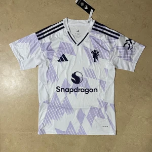 Manchester United tröja - Snygg Manchester United fotbollströja från Adidas med Snapdragon sponsortryck. Tröjan är vit med ljuslila geometriskt mönster, har svarta Adidas-ränder på axlarna och klubbmärke på bröstet. Tillverkad i lätt och ventilerande material, perfekt för träning eller match.