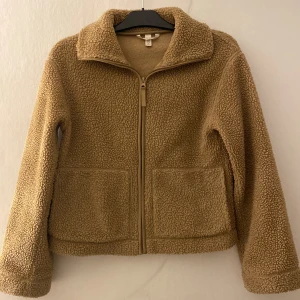 Beige teddyjacka från H&M XS - Mysig beige teddyjacka från H&M i storlek XS. Jackan har dragkedja framtill, två stora fickor och en klassisk krage. Perfekt för kyliga dagar med sin fluffiga och trendiga look.