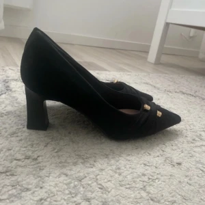 Svarta klackar med spetsig tå och gulddetaljer - Stilrena svarta pumps från NA-KD i mocka-look med spetsig tå och låg klack. Skorna har snygga guldfärgade detaljer framtill som ger en extra touch. Perfekta för dig som gillar klassisk men trendig stil.