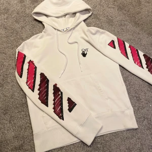 Vit Off-White hoodie  - Säljer en vit hoodie från Off-White med ikoniska grafiska röda och svarta tryck på ärmar och rygg. Tröjan har huva med snörning och en stor magficka. Perfekt statement-plagg för dig som gillar streetwear och unika prints.