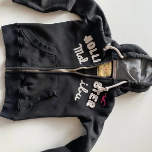 Svart hoodie från Hollister  - Svart hoodie från Hollister med vit broderad text på bröstet. Tröjan har dragkedja, stora fickor fram, vit snörning i huvan och grått innerfoder. Perfekt för chill dagar och snygg streetstil. Tröjan är liten i storlek skulle uppskatta storlek (S) men det står (M) 