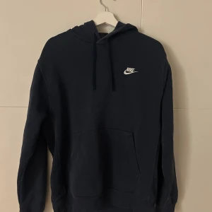 Mörkblå hoodie från Nike med huva - Mörkblå hoodie från Nike med klassisk logga i vitt på bröstet. Tröjan har huva med snörning, stor magficka och långa ärmar. Perfekt för chill dagar och streetstyle. Mjuk bomullskänsla och relaxed passform.
