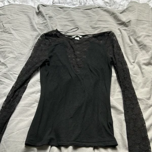Svart spetsblus med v-ringning H&M - Svart långärmad blus från H&M med djup v-ringning och transparenta spetsärmar. Toppen har en figurnära passform och är tillverkad i ett mjukt material med spetsdetaljer upptill och på ärmarna. 