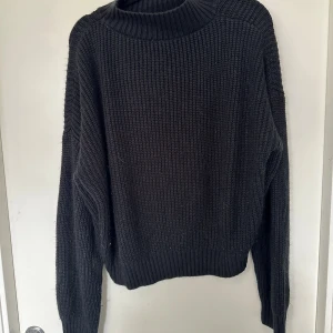 Svart stickad tröja med ribbad krage S-M - En svart stickad tröja med grov struktur och ribbad hög krage. Tröjan har långa ärmar och ribbade muddar vid ärmslut och nederkant. Perfekt för lager-på-lager och chill höst- och vinterstil.