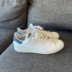 Adidas Stan Smith vita sneakers strl 43 1/3 - Klassiska Adidas Stan Smith sneakers i vitt med mörkblå detalj på hälen. Använda fåtal gånger som nya. Skick 9/10. Hör av dig vid frågor!