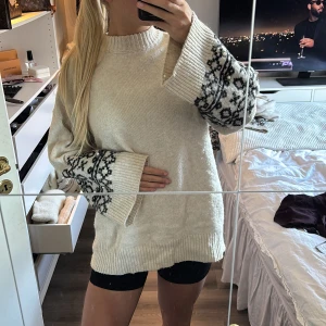 Beige stickad tröja med mönstrade ärmar - Säljer en oversized beige stickad tröja med breda långärmade muddar som har svart mönster och pärldetaljer. Tröjan har ribbad hals och nederkant, och är perfekt för dig som gillar mysiga och unika plagg med lite extra detaljer på ärmarna.