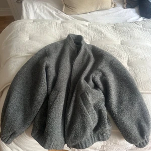 Grå teddyjacka med dragkedja - Säljer en oversized grå teddyjacka med grov struktur och hög krage. Jackan har långa ärmar med resår vid muddar och två snedställda fickor framtill. Stängs med dragkedja och har en chill, loose passform som är perfekt för kalla dagar.