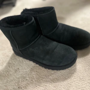 Svarta UGGs - Svarta UGGs i klassisk modell med rund tå och platt sula. Tillverkade i mjuk mocka med fluffigt foder på insidan för extra värme. Perfekta för kalla dagar och riktigt sköna att dra på sig när det är kyligt ute. Jag har använt dom en vinter och dom har varit super bra! Det är storlek 42 men passar som en 43 
