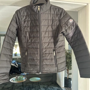 Svart quiltad dunjacka från Moncler - Snygg svart dunjacka från Moncler med quiltad design och hög krage. Jackan har dragkedja framtill, två fickor med dragkedja och Moncler-logga på ärmen. Perfekt för kyliga dagar och ger en clean, stilren look.