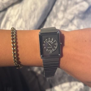 Omega Seamaster fyrkantig klocka silver - Snygg Omega Seamaster med fyrkantig svart urtavla och datumvisning. Klockan har en stilren länk i silverfärgat stål och klassisk Omega-logga på spännet. Klockan är från 70-80 tal, den är lite repig och behöver byta batteri. 