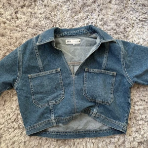 Croppad jeansjacka från Zara - Säljer en croppad jeansjacka från Zara i klassisk blå denim. Jackan har två stora fickor framtill, krage och en halv dragkedja.