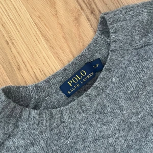Grå stickad tröja Polo Ralph Lauren - Grå stickad tröja från Polo Ralph Lauren med rund hals och ribbade muddar. Tröjan har snygga bruna mockadetaljer på armbågarna som ger en cool touch. Perfekt för dig som gillar klassisk stil med en twist.
