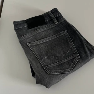 Grå jeans från Jack & Jones - Säljer ett par grå jeans från Jack & Jones med snyggt slitna detaljer och klassisk femficksdesign. Jeansen har raka ben och subtila sömmar på bakfickan. Perfekta för dig som gillar en avslappnad men stilren look. De har en liten defekt på högra ficken av en mobil, Storleken är W31 L32 och tveka inte på att kontakta mig vid mer information!