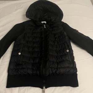 Svart cardigan från Moncler - Snygg svart cardigan från Moncler med huva och dragkedja. Den har långa ärmar och ribbade muddar. Den är quiltad och har ett Moncler-märke på ärmen. Perfek för hösten/våren priset kan diskuteras 
