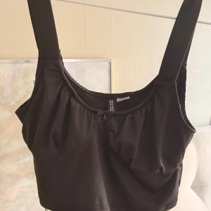 Svart croppad linnetopp från Divided - Svart croppad linnetopp från Divided med breda axelband och lätt rynkad detalj vid bysten. Toppen har en enkel och stilren design som passar perfekt till jeans eller kjol. Mjuk och stretchig känsla.