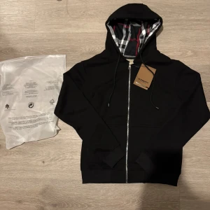 Svart Burberry hoodie med dragkedja - Svart hoodie från Burberry med klassiskt rutigt foder i huvan och silverfärgad dragkedja. Tröjan har snörning i huvan, ribbade muddar och är tillverkad i mjuk bomull. Perfekt för dig som gillar stilrena och exklusiva plagg med ikoniska detaljer.