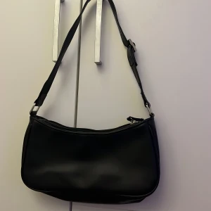 Svart axelväska från Monki - Snygg svart axelväska från Monki i stilren design. Väskan är tillverkad i slätt syntetmaterial och har justerbart axelband samt dragkedja upptill. Perfekt storlek för det viktigaste och passar till många olika outfits.