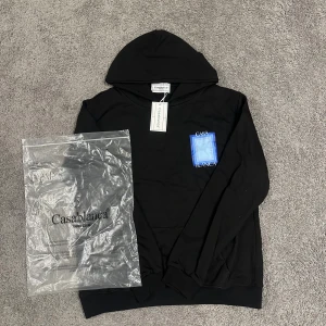 Svart hoodie från Casablanca - Svart hoodie från Casablanca med stor blå och vit grafisk ruta och texten 'CASABLANCA' på både bröstet och ryggen. Tröjan har huva och ribbade muddar. Tillverkad i mjuk bomull, perfekt för en chill och stilren look.