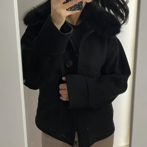 Svart pälsjacka från Neo Noir - Säljer en svart pälsjacka från NOIR med fluffig fuskpäls runt kragen och stora fickor framtill. Jackan har en croppad passform, knappar och långa ärmar. Perfekt för dig som vill ha en cool och stilren look under höst och vinter.