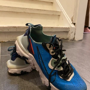 Nike React Element 87 blå sneakers - Nike React Element 87 sneakers i blått med svarta och vita detaljer samt inslag av mintgrönt och orange. Skorna har en genomskinlig mesh-överdel, svart snörning och en chunky vit sula med svarta partier. Snygg och futuristisk design med bekväm passform och coola detaljer. Utan kartong, går ner i pris vid snabb affär 