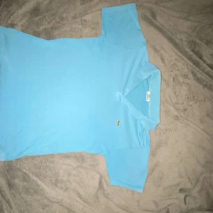 Ljusblå pikétröja från Lacoste - Snygg ljusblå pikétröja från Lacoste med klassisk krage och knappar framtill. Tröjan har det ikoniska krokodilmärket broderat på bröstet och är tillverkad i mjuk bomull. Perfekt för en fräsch och sportig look.