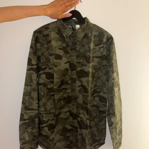 Grön camo skjorta från H&M, stl M - Säljer en grön långärmad skjorta från H&M i storlek M. Skjortan har camouflage-mönster med blommiga detaljer och klassisk krage. Tillverkad i bomull och har knäppning framtill samt vid ärmslut. Perfekt för dig som gillar streetstyle och unika prints.