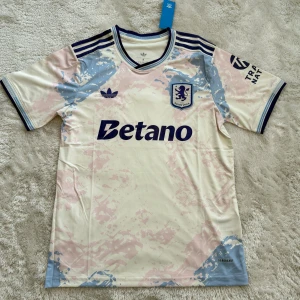 Aston Villa Adidas fotbollströja - Snygg Aston Villa fotbollströja från Adidas med Betano-tryck på bröstet. Tröjan är kortärmad och har en ljus bas med blå och rosa mönsterdetaljer samt mörkblå ränder på axlarna. Tillverkad i lätt och ventilerande material, perfekt för match eller träning.