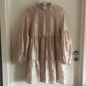 Beige volangklänning från Gina Tricot - Beige kortklänning från Gina Tricot med volanger och ballongärmar. Klänningen har hög hals, knappar i nacken och är tillverkad i ett glansigt material som ger en lyxig känsla. 