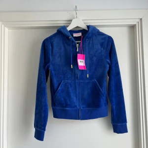 Blå velour hoodie Juicy Couture XS - Juicy Couture hoodie i en snygg, klarblå velour. Tröjan har huva, dragkedja framtill, snörning med gulddetaljer och två fickor. Perfekt för en chill och trendig look. Broderad logga på bröstet och ribbade muddar vid ärmslut och midja.