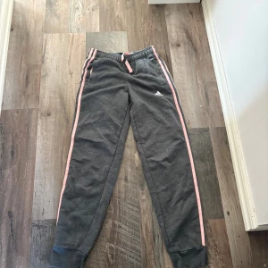 Grå Adidas mjukisbyxor med rosa detaljer - Grå mjukisbyxor från Adidas med rosa ränder längs sidorna och matchande rosa snörning i midjan. Byxorna har resår och muddar vid bensluten samt Adidas-logga på låret. Perfekta för chill eller träning.