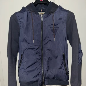 Blå hybridjacka Aeronautica Militare - Snygg blå vindjacka från Aeronautica Militare med avtagbar huva och dubbelzip framtill. Jackan har ribbade muddar, två fickor med dragkedja. Schyssta armbågspatches och huvsnören. Italiensk strl - S mer som XS.