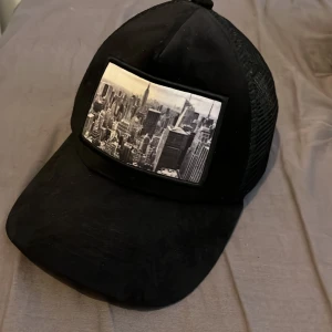 Svart velvet keps från Gårda - Svart truckerkeps från Gårda med mesh på baksidan och justerbar snapback. Framtill finns en stor svartvit patch med stadsmotiv. Kepsen har klassisk böjd skärm och ventilerande nätmaterial bak, perfekt för en cool streetstil.