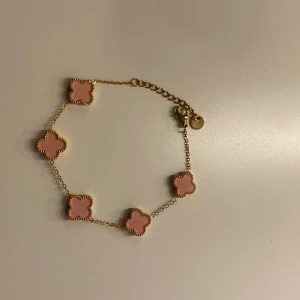 Guldfärgat armband med rosa klöver - Superfint armband i guldfärgat rostfritt stål med fem rosa fyrklöverformade hängen. Kedjan är tunn och smidig med justerbar längd och en liten rund bricka med märkning. Perfekt för dig som gillar söta detaljer och stilren design.