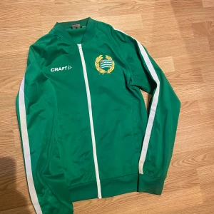  Hammarby ziptröja - Grön ziptröja från Craft med Hammarby IF-logga på bröstet och vita ränder längs ärmarna. Står 'HAMMARBY' på ryggen. Köpt på skrapan för 750.