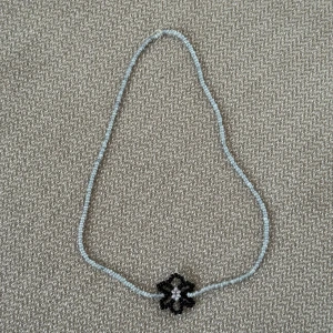 Pärlhalsband med svart blomma - Handgjort halsband med små ljusblå pärlor och en söt blomma i svart och vitt i mitten. Perfekt accessoar för att ge din outfit en unik och personlig touch. Det är 45cm långt 🩶🩵